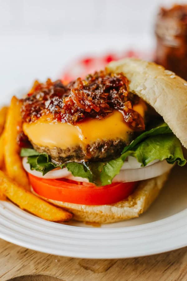 Close-up of a cheeseburger with lettuce, tomato, onion, cheddar cheese, and a generous topping of caramelized onion jam, served on a sesame bun with French fries on a white plate.