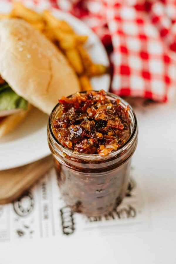 A jar of chunky bacon jam sits on a table beside a plate with a burger and fries. A red and white checkered cloth is in the background.