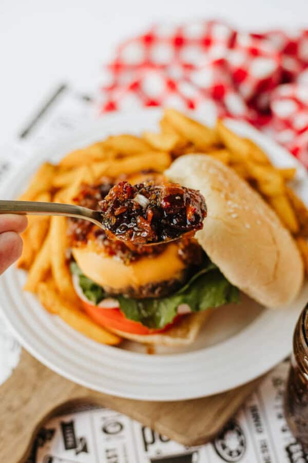 A hand holds a spoonful of relish or chutney above a cheeseburger with lettuce and tomato, served with French fries on a white plate. A red and white checked cloth is in the background.