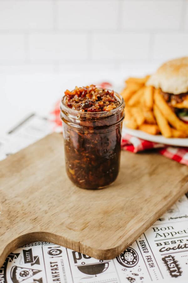 A glass jar filled with bacon jam sits on a wooden cutting board. In the background, there is a plate with a burger and French fries. The scene appears to be set on a table with food-themed paper.