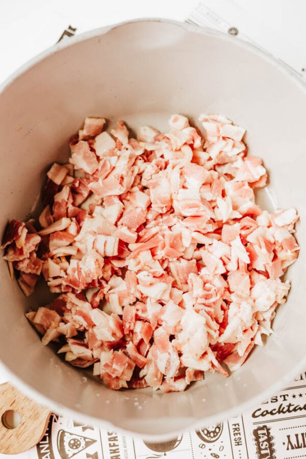 A pot filled with chopped raw bacon pieces sitting on a countertop, with part of a patterned paper or placemat visible underneath.