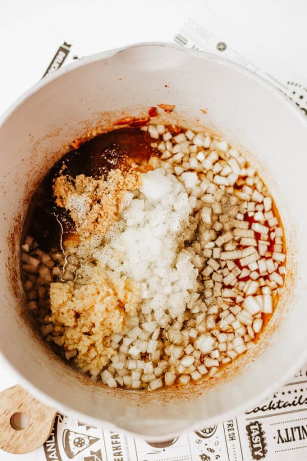 Chopped onions, minced garlic, brown sugar, and sauces in a white pot, ready to be mixed, sitting on top of printed paper.