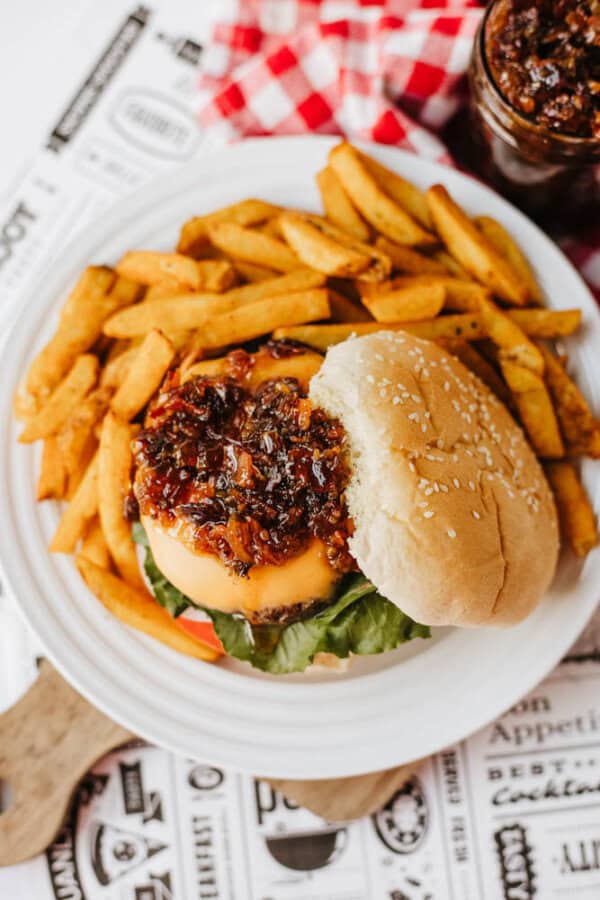 A cheeseburger topped with caramelized onions on a sesame bun, served with a side of thick-cut French fries on a white plate, placed on a decorative paper mat with a red and white checkered napkin nearby.