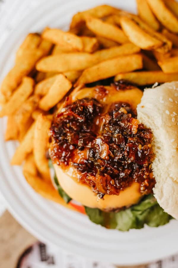 A close-up of a cheeseburger topped with caramelized onions, lettuce, and tomato on a sesame seed bun, served with a generous portion of golden French fries on a white plate.
