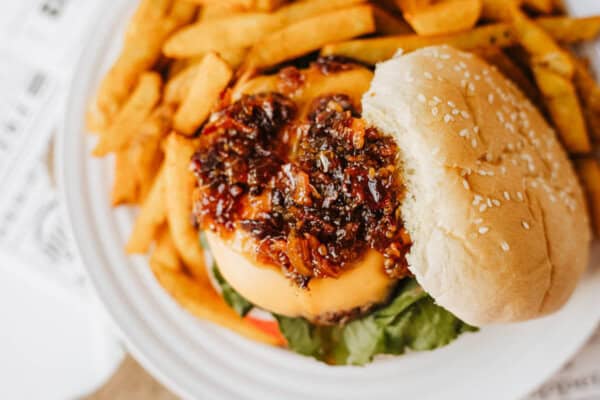 A close-up of a cheeseburger topped with caramelized onions on a sesame seed bun, with lettuce and tomato, served alongside a pile of golden French fries on a white plate.