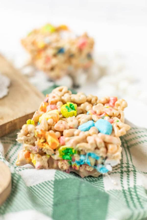 A close-up of a colorful cereal treat bar with pieces of cereal and marshmallows, resting on a green and white checkered cloth. Another treat bar is blurred in the background.