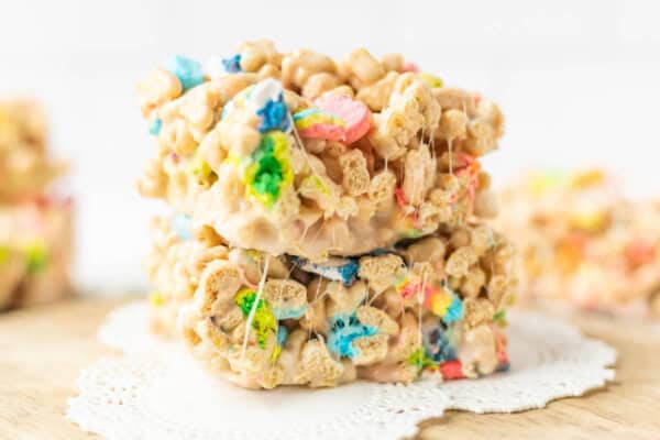 Two stacked marshmallow cereal treats with colorful bits, likely made from a marshmallow and multicolored cereal mix, sit on a white doily atop a wooden surface with more treats blurred in the background.