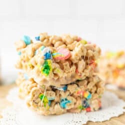Two stacked cereal treats made with colorful marshmallow cereal and melted marshmallow, placed on a wooden board with a decorative doily.