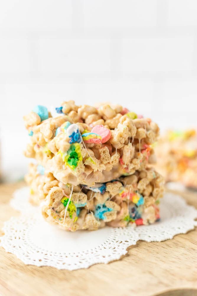Two stacked cereal treats made with colorful marshmallow cereal and melted marshmallow, placed on a wooden board with a decorative doily.