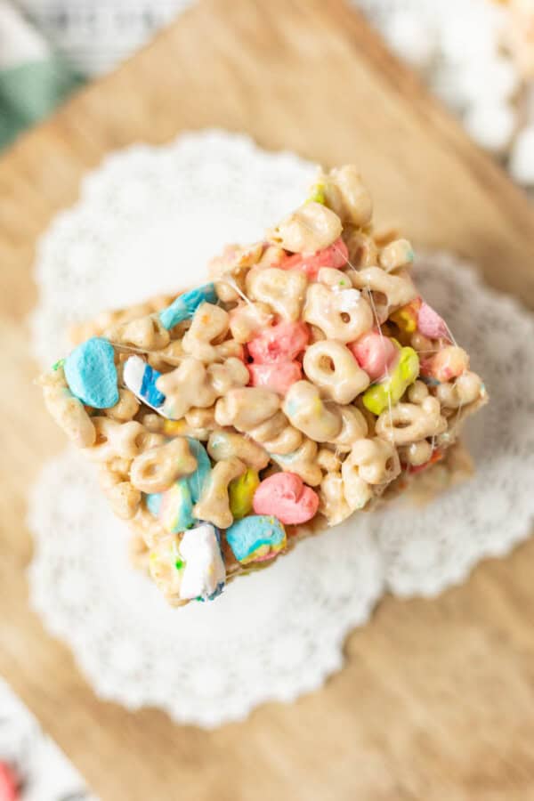 A square cereal treat made with colorful marshmallow pieces and oat cereal, resting on a white paper doily atop a wooden surface.