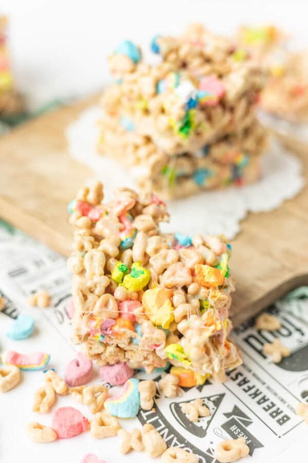 A close-up of a colorful dessert bar made from Lucky Charms cereal and marshmallows, with cereal pieces and marshmallows scattered around, set on a wooden board and newspaper-style surface.
