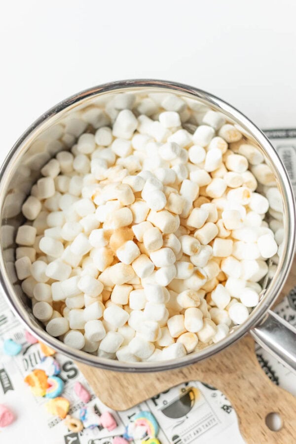 A saucepan filled with mini marshmallows, some lightly toasted, sits on a wooden cutting board with colorful candy pieces nearby.