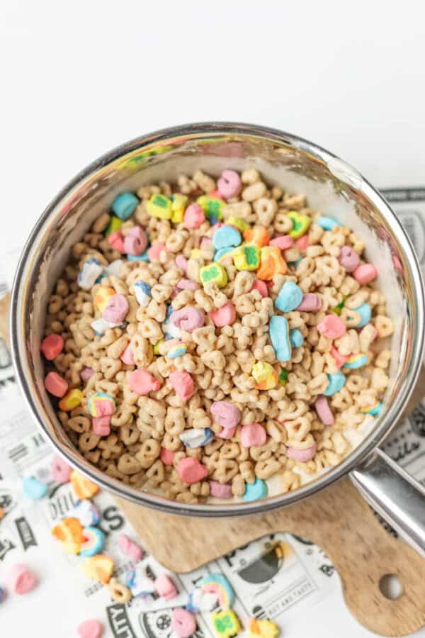A saucepan filled with colorful marshmallows and round cereal pieces sits on a wooden board, with some cereal and marshmallows scattered nearby.