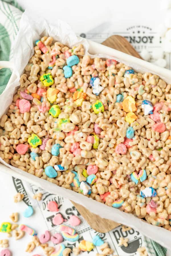 A tray of cereal bars made with colorful marshmallows and round oat cereal pieces, resting on white parchment paper. Some cereal pieces and marshmallows are scattered nearby.