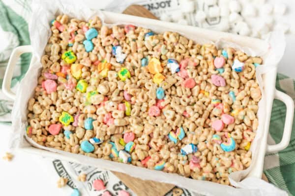A rectangular baking dish lined with parchment paper is filled with a colorful cereal treat made of Lucky Charms cereal and marshmallows, set on a light cloth with mini marshmallows nearby.