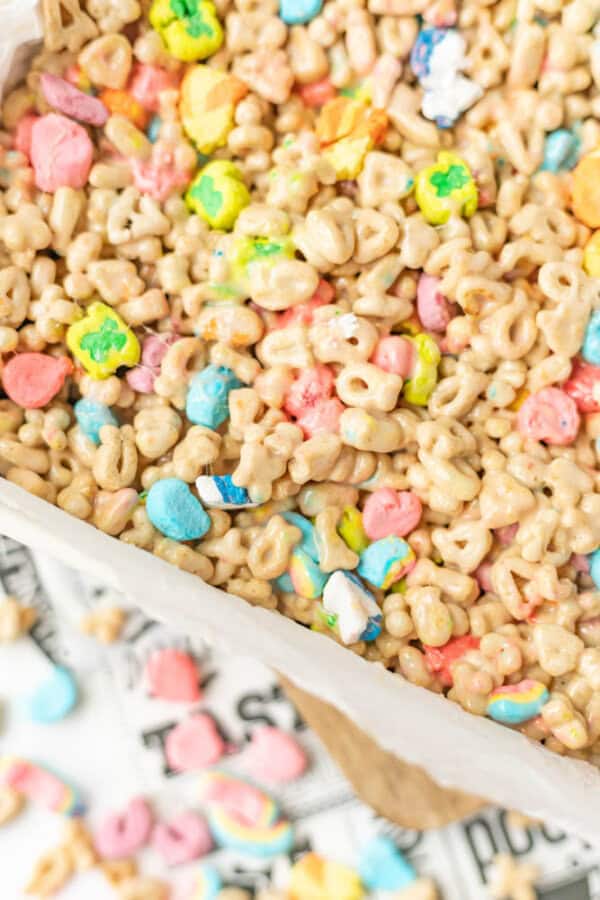 A close-up of a colorful cereal mix featuring marshmallow shapes and glazed oat pieces, spread in a lined baking dish. Some marshmallows and cereal pieces are scattered on a newspaper beneath the dish.