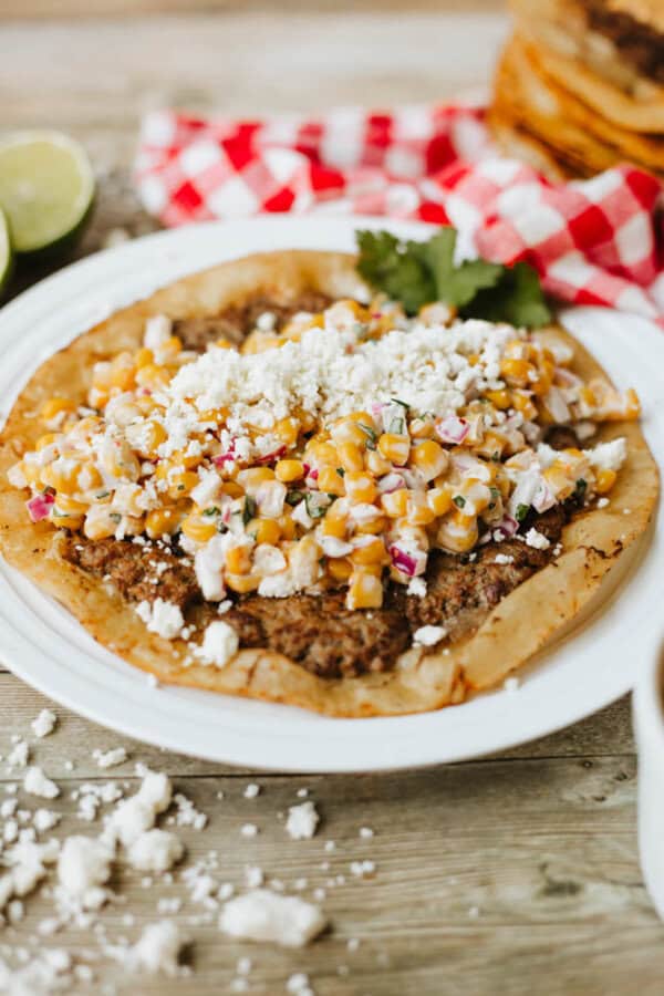 A close-up of a tlayuda topped with refried beans, corn salad, chopped red onion, cilantro, and crumbled cheese on a white plate, with a red plaid cloth and lime halves in the background.