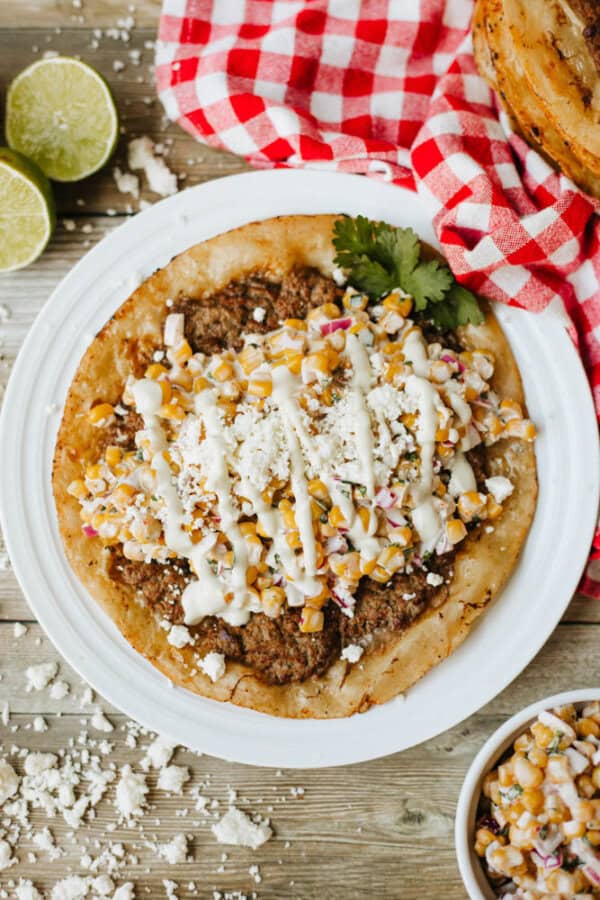A tostada topped with seasoned ground meat, corn salsa, crumbled cheese, and a drizzle of crema sits on a white plate. A red checkered cloth, lime halves, and cheese crumbles surround the plate.
