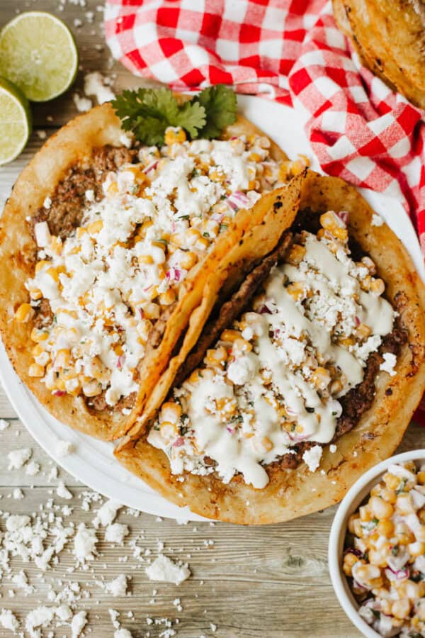 Two Mexican-style tostadas topped with refried beans, corn, cheese, and creamy sauce, served on a white plate with lime wedges and a red checkered napkin in the background.