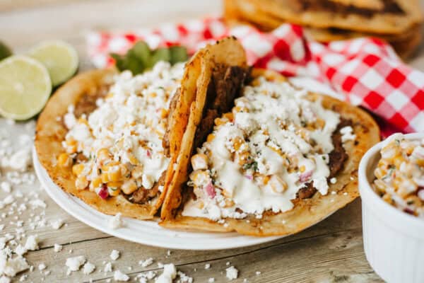 Two tacos filled with corn, diced vegetables, crumbled cheese, and creamy sauce on a plate, with a side of lime slices and a red-checkered napkin in the background.