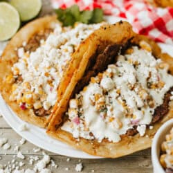 Two Mexican-style tostadas topped with creamy corn, black beans, and crumbled cheese are served on a white plate. Lime halves and a red-checkered cloth are in the background. Crumbled cheese is scattered on the table.