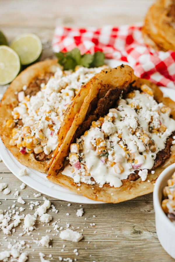 Two Mexican-style tostadas topped with creamy corn, black beans, and crumbled cheese are served on a white plate. Lime halves and a red-checkered cloth are in the background. Crumbled cheese is scattered on the table.