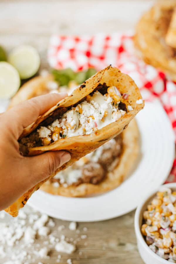 A hand holds a taco with seasoned meat, corn, cheese, and cream sauce. A plate with a similar taco, a bowl of corn topping, and lime wedges are visible in the background.