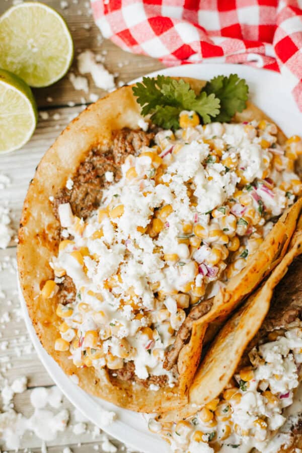 Two tacos on a white plate, topped with seasoned meat, corn salsa, crumbled white cheese, fresh cilantro, and creamy sauce. Lime halves and a red checkered napkin are in the background.