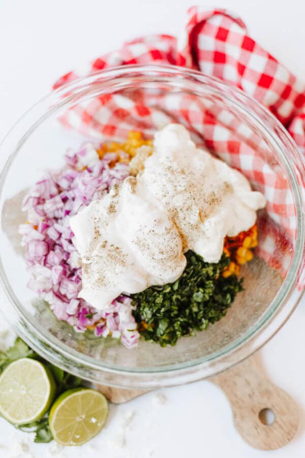 A glass bowl filled with chopped red onion, leafy herbs, corn, and a dollop of creamy sauce topped with black pepper. Next to the bowl are sliced limes on a wooden board and a red checkered cloth.