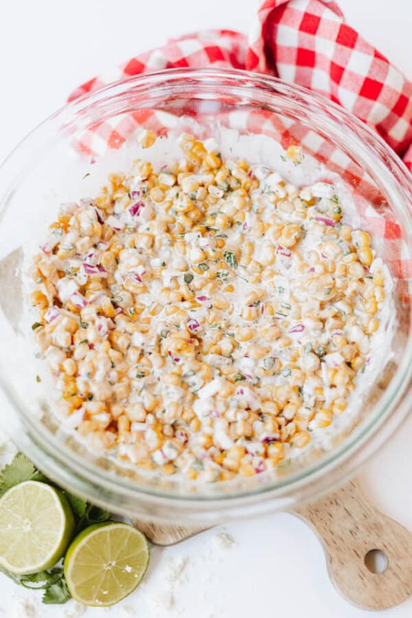 A glass bowl filled with creamy corn salad containing corn, chopped red onions, and herbs, sits on a wooden board. Beside it are lime halves and a red-and-white checkered cloth.