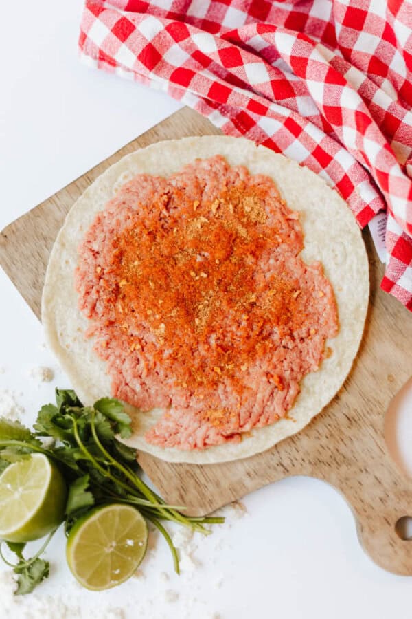 A tortilla topped with ground meat and orange seasoning sits on a wooden board. Nearby are cilantro, halved limes, scattered flour, and a red-and-white checkered cloth.