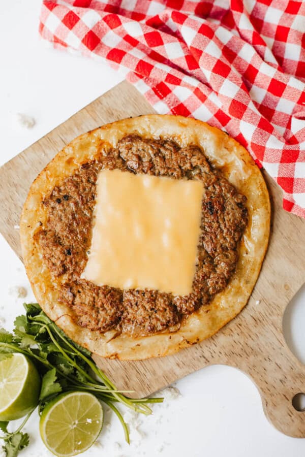A flatbread topped with minced meat and a slice of melted cheese sits on a wooden board next to a bunch of cilantro, halved limes, and a red-and-white checkered cloth.