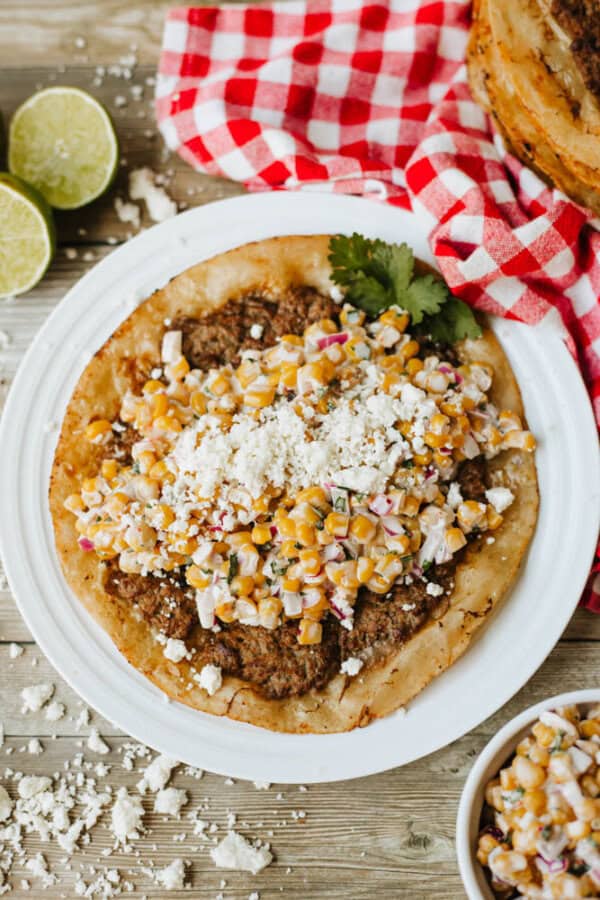A white plate with a tortilla topped with seasoned meat, corn salad, crumbled cheese, and cilantro sits on a wooden surface next to lime halves and a red checkered cloth.