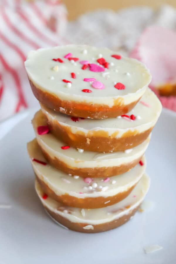 A stack of four round, layered white and caramel-colored candies topped with white, pink, and red sprinkles, placed on a white plate with a soft, striped cloth in the background.
