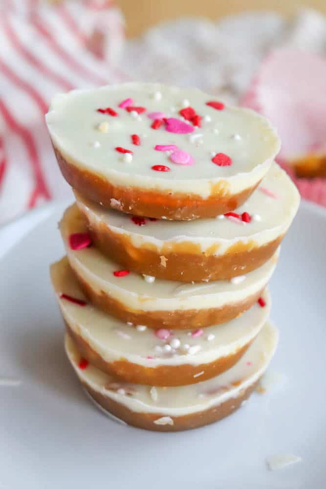 A stack of four round, layered white and caramel-colored candies topped with white, pink, and red sprinkles, placed on a white plate with a soft, striped cloth in the background.