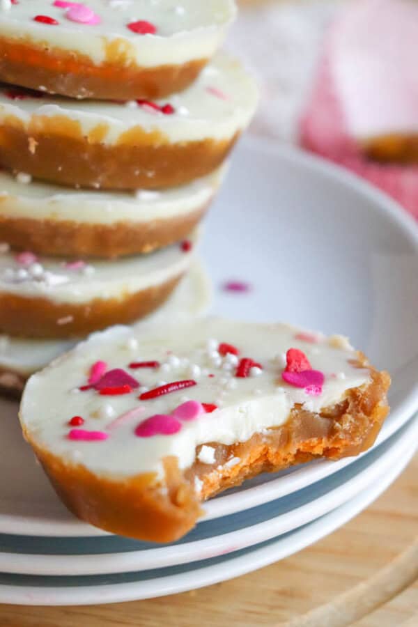 A close-up of white chocolate peanut butter cups with heart-shaped pink and red sprinkles, stacked on white plates. One cup in the foreground has a bite taken out, revealing a creamy peanut butter layer inside.