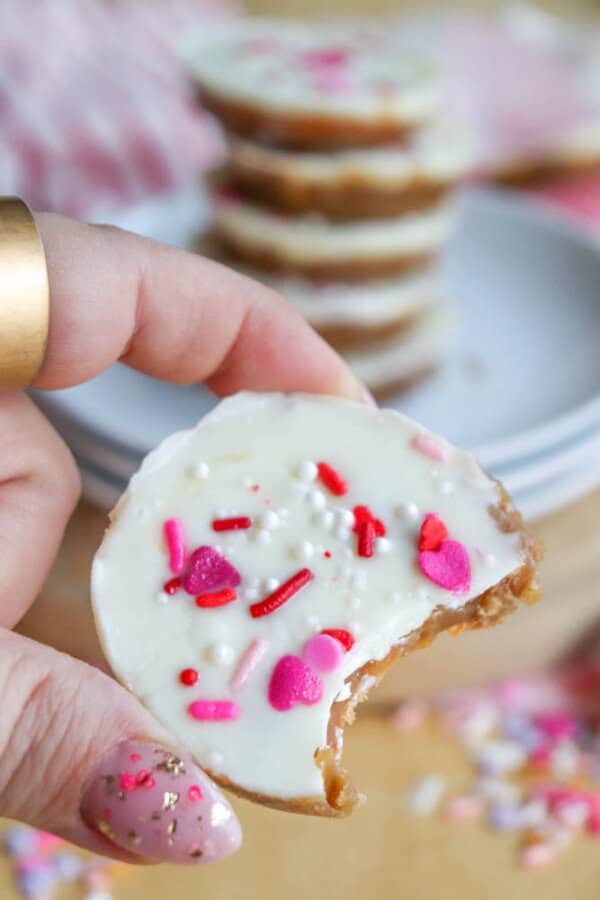A hand holds a round, bite-sized treat topped with white icing and pink, red, and white heart-shaped sprinkles. Stacked treats and more sprinkles are blurred in the background.