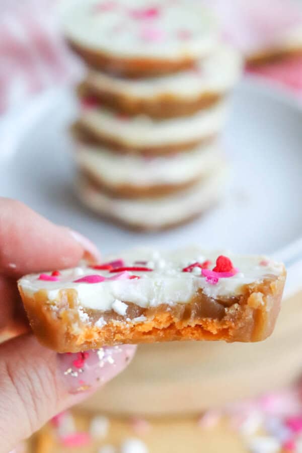 A hand holding a partially eaten round treat with a caramel base, white chocolate topping, and pink heart sprinkles. A blurred stack of similar treats sits on a plate in the background.