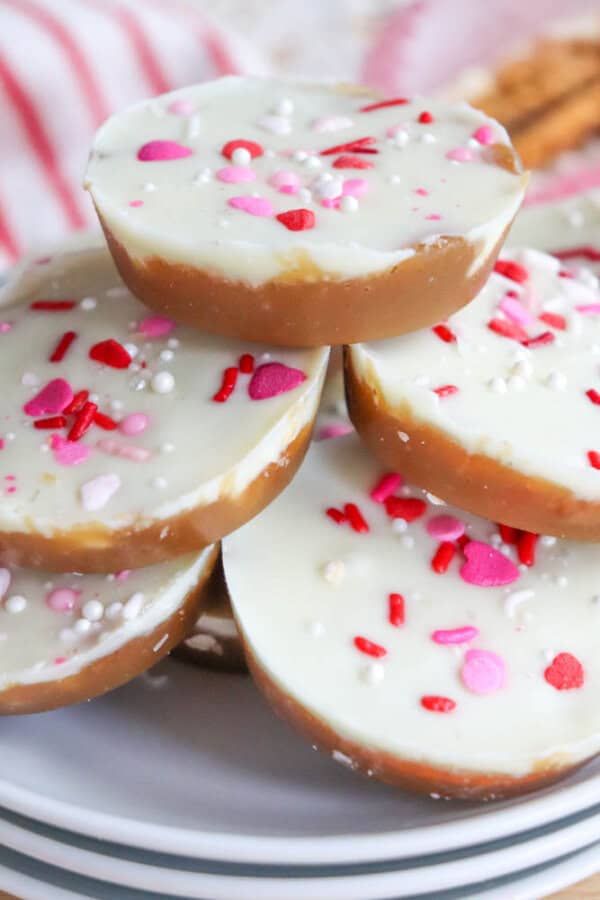A stack of round, layered treats with a caramel base and white chocolate topping, decorated with pink, red, and white heart-shaped and round sprinkles, arranged on a white plate.