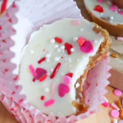 A white chocolate peanut butter cup with colorful heart and sprinkle toppings, wrapped in a pink striped liner, sits on a wooden surface surrounded by scattered sprinkles. A bite has been taken from the treat.