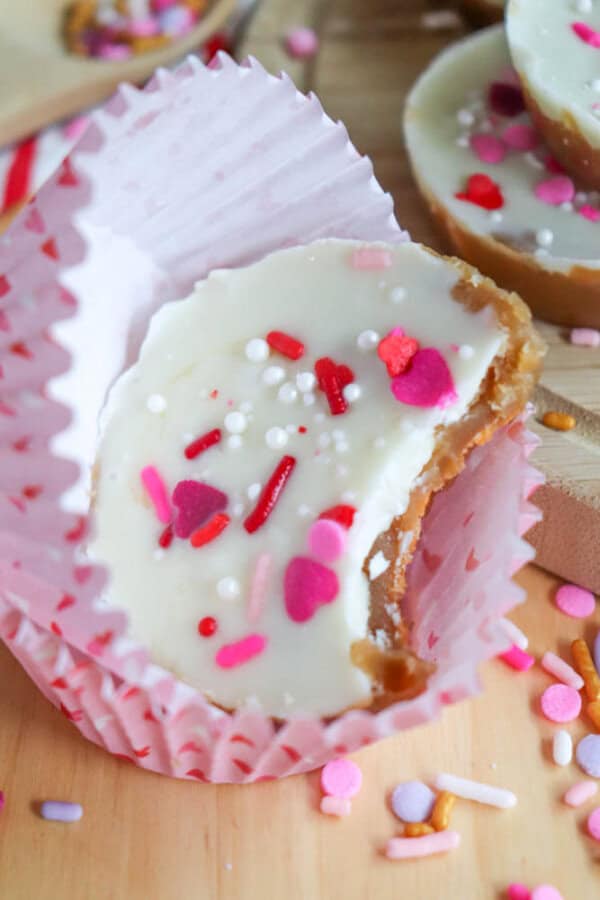 A white chocolate peanut butter cup with colorful heart and sprinkle toppings, wrapped in a pink striped liner, sits on a wooden surface surrounded by scattered sprinkles. A bite has been taken from the treat.