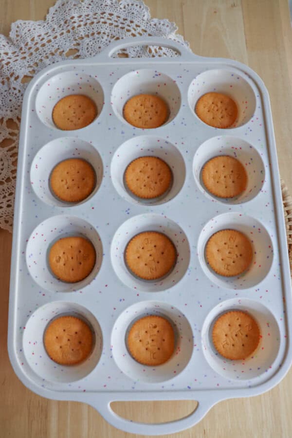 A white muffin tray with 12 round wells, each containing a single plain biscuit at the bottom. The tray sits on a wooden surface with a lacy white cloth nearby.