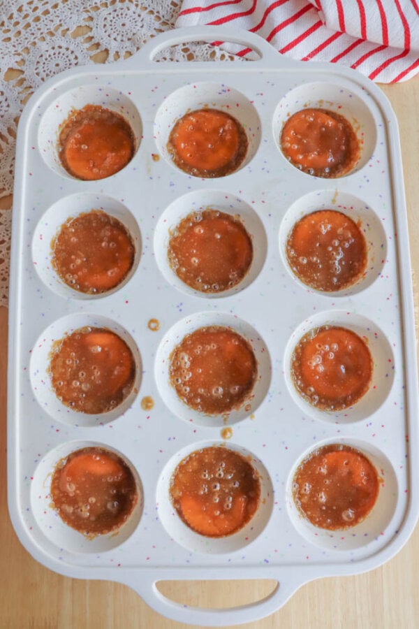 A white muffin pan filled with brown bubbly liquid in each cup, sitting on a light wood surface. A red-striped towel and lace fabric are partially visible in the background.