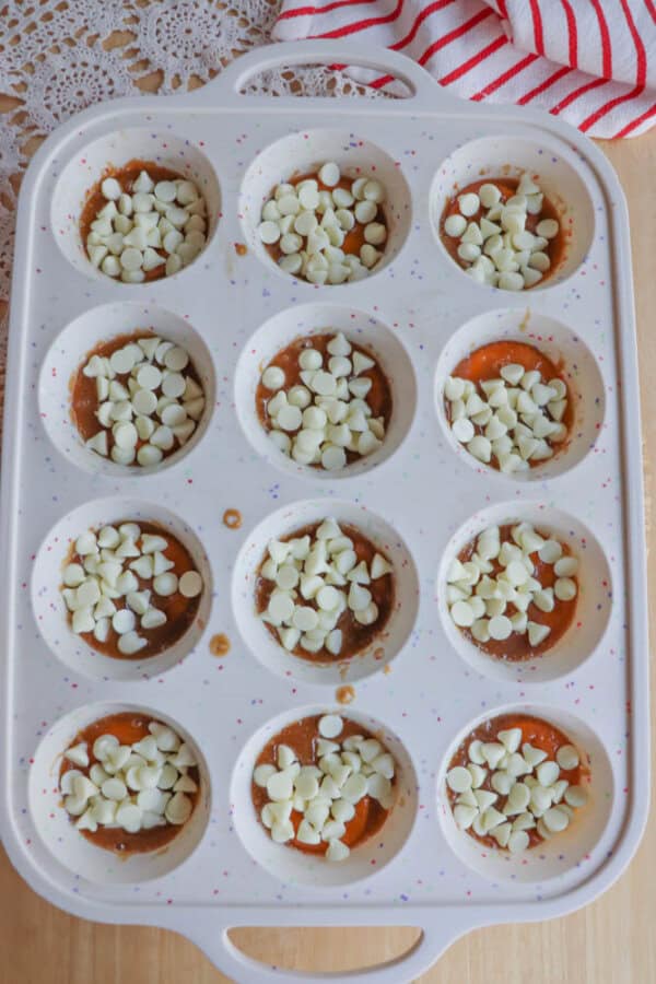 A white 12-cup muffin pan filled with partially baked batter, topped with white chocolate chips. The pan sits on a wooden surface near a red-striped towel and lace fabric.