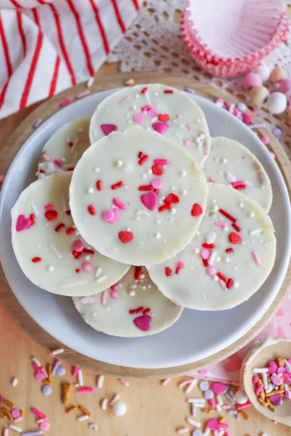 White chocolate bark circles topped with pink, red, and white heart and sprinkle decorations are arranged on a white plate. A striped towel, cupcake liners, and scattered sprinkles are in the background.