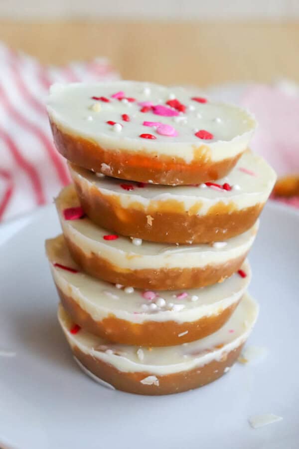 A stack of round layered treats with a white chocolate top, caramel-colored base, and decorated with pink, red, and white sprinkles, placed on a white plate.