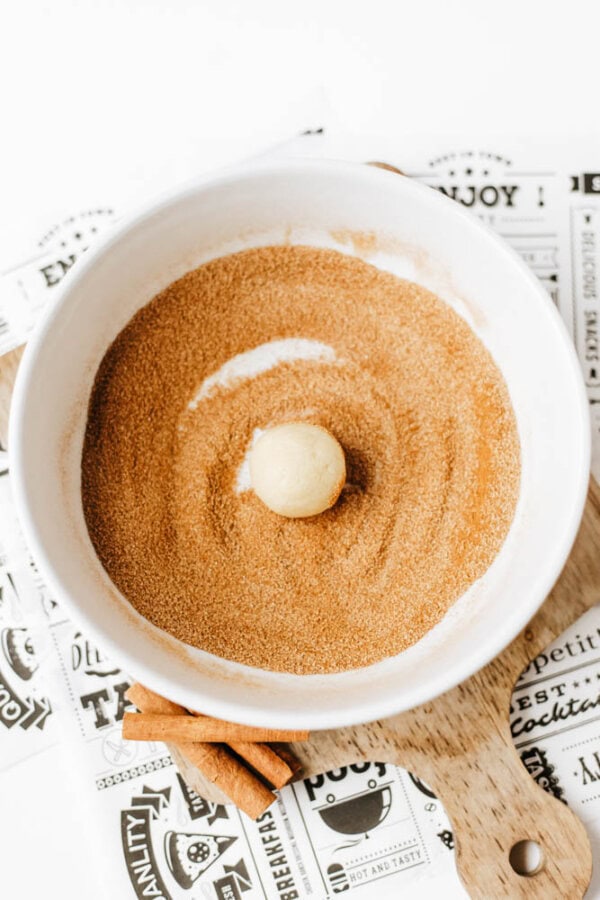 A ball of dough sits in the center of a white bowl filled with cinnamon sugar, ready to be rolled. The bowl is placed on a wooden board with printed paper and cinnamon sticks nearby.