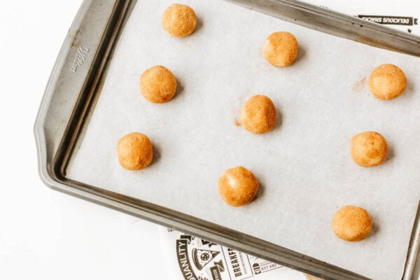 A baking sheet lined with parchment paper holds eight evenly spaced, round cookie dough balls coated in cinnamon sugar, ready to be baked.