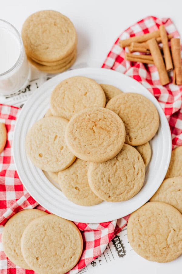 A plate of round sugar cookies sits on a red and white checkered cloth, with more cookies nearby. A glass of milk and cinnamon sticks are next to the plate on a white surface.