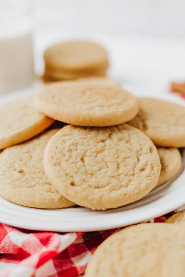 A plate of golden brown sugar cookies stacked on top of each other, placed on a red and white checkered cloth with a glass of milk and more cookies blurred in the background.